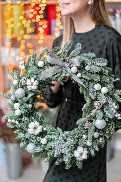 Het winkelen van Kerstmis. Groene Kerst krans in vrouwelijke handen. Versierd met grijze elementen, katoen en zilveren bal. Garland bokeh op achtergrond. — Stockfoto