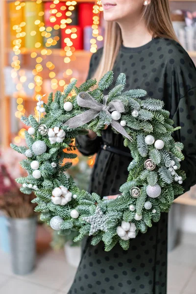 Χριστουγεννιάτικα ψώνια. Πράσινο Χριστουγεννιάτικο στεφάνι στα γυναικεία χέρια. Διακοσμημένο με γκρι ΣΤΟΙΧΕΙΑ της συνθεσης, βαμβάκι και ασημένια σφαίρα. Γιρλάντα bokeh στο παρασκήνιο. — Φωτογραφία Αρχείου