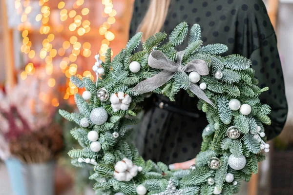 Compras de Navidad. Corona de Navidad verde en manos femeninas. Decorado con elementos grises, algodón y bola de plata. Guirnalda bokeh en el fondo . — Foto de Stock