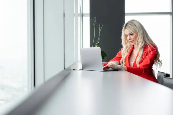 Kırmızı mercan iş takım elbiseli kadın. Sarışın genç iş kadını ile modern gri laptop pencerenin önünde yüksek kat ofisinde. Kahve içme kız — Stok fotoğraf
