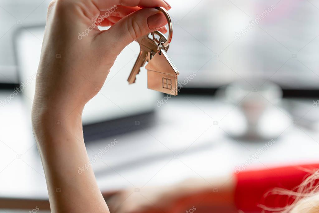 Mortgage concept. Woman in red coral business suit holding key with house shaped keychain. Modern light lobby interior. Real estate, hypothec, moving home or renting property.