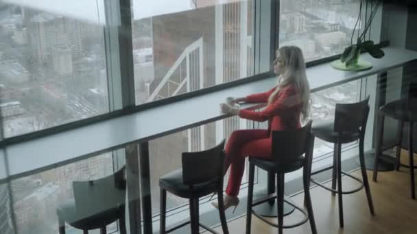 Mujer en traje de negocios de coral rojo bebe café en una mesa alta cerca de la ventana. Joven mujer de negocios rubia en la oficina en el piso alto delante de la ventana . — Vídeos de Stock