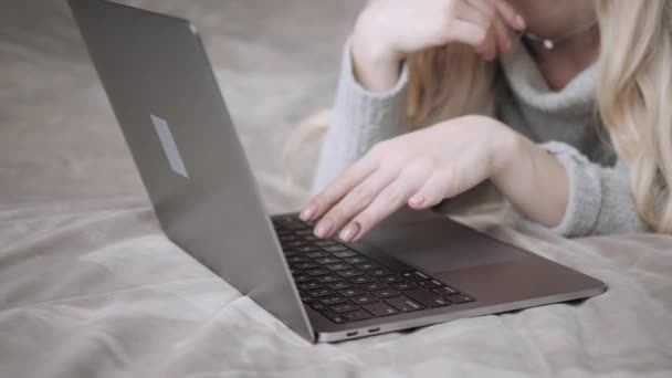 Bella donna bionda sdraiata sul letto con computer portatile in alluminio grigio. La ragazza sorride, di buon umore. Blogging, navigazione internet, chat. In un maglione accogliente caldo e calzini di lana. Buon inverno concetto . — Video Stock