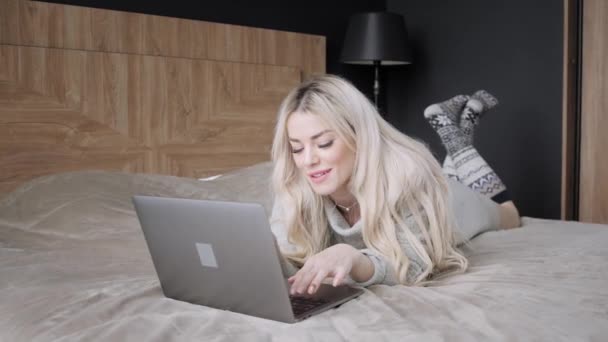 美丽的金发碧眼的女人躺在床上与灰色的铝笔记本电脑。女孩微笑, 心情好。写博客, 浏览互联网, 聊天。穿着温暖舒适的毛衣和羊毛袜子。快乐的冬天概念. — 图库视频影像