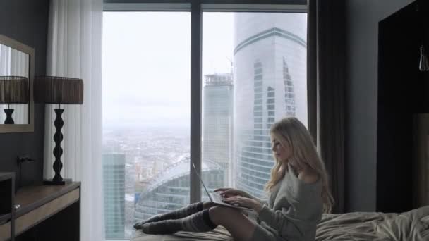 Красива блондинка сидить на ліжку з сірим алюмінієвим ноутбуком. Дівчина посміхається, гарний настрій. Блог, перегляд інтернету, балачки. У теплому затишному светрі і вовняних шкарпетках. Щаслива зимова концепція . — стокове відео