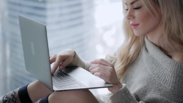 Mooie blonde vrouw zittend op het bed met grijs aluminium laptop. Meisje glimlacht, goed humeur. Bloggen, surfen op internet, chatten. In een warme, knusse trui en wollen sokken. Happy winter concept. — Stockvideo