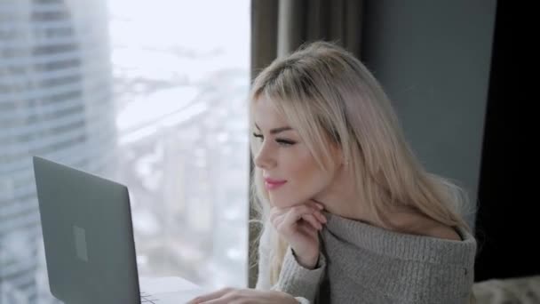Όμορφη ξανθιά γυναίκα που κάθεται στο κρεβάτι με το φορητό υπολογιστή από αλουμίνιο γκρι. Κορίτσι χαμόγελα, καλή διάθεση. Blogging, περιήγηση στο internet, κουβεντιάζοντας. Σε έναν ζεστό άνετο πουλόβερ και κάλτσες μαλλιού. Καλό χειμώνα έννοια. — Αρχείο Βίντεο