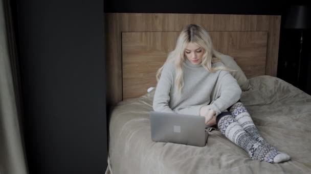 Красива блондинка сидить на ліжку з сірим алюмінієвим ноутбуком. Дівчина посміхається, гарний настрій. Блог, перегляд інтернету, балачки. У теплому затишному светрі і вовняних шкарпетках. Щаслива зимова концепція . — стокове відео