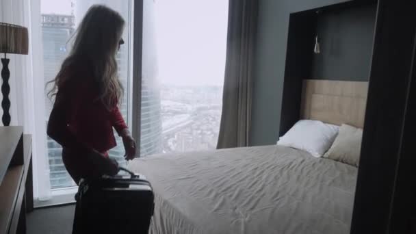 Joven mujer de negocios rubia llega a una habitación de hotel con maleta negra. Mujer con traje de negocios de coral rojo. Chica joven entra en la habitación sosteniendo la maleta por el mango — Vídeo de stock