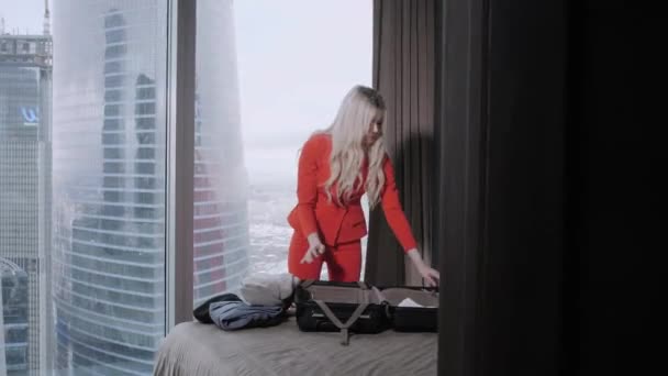 Die junge blonde Geschäftsfrau kommt mit schwarzem Koffer in ein Hotelzimmer. Frau im Business-Anzug aus roten Korallen. junges Mädchen packt Koffer aus. — Stockvideo