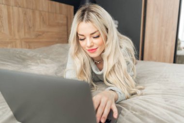bir laptop perde arkasında bir kız portresi. Güzel sarışın kadın yatakta yatıyordu ve dizüstü bilgisayar ekranında görünüyor. Blogging, internet tarama, sohbet. Bir sıcak rahat kazak
