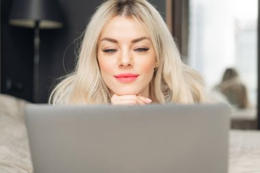 bir laptop perde arkasında bir kız portresi. Güzel sarışın kadın yatakta yatıyordu ve dizüstü bilgisayar ekranında görünüyor. Blogging, internet tarama, sohbet. Bir sıcak rahat kazak