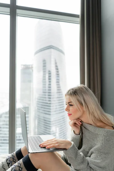 stock image Beautiful blonde woman sitting on the bed and looks at the laptop screen.. Girl smiles, good mood. Blogging, browsing internet, chatting. In a warm cozy sweater and wool socks. Happy winter concept.