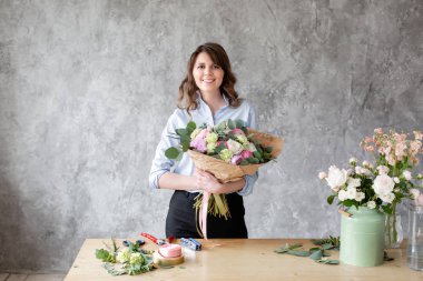 Genç gülümseyen kadın çiçek sanatçı kameraya, arayan samimi lüks bir buket elinde tutar. Profesyonel çiçekçi bir buket pembe Düğünçiçeği ve peonies bir çiçekçide yapma.
