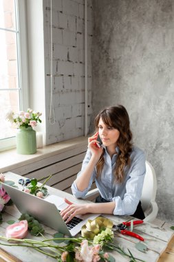 Genç dostu kadın çiçekçi bir cep telefonu üzerinde konuşurken gülümseyen ve bir dizüstü bilgisayarda temel çalışma sipariş oluşturun. Çiçek teslimat kavramı. Bir çiçekçi dükkanı profesyonel işçi.