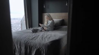 Akıllı telefon ile yatağa oturan rüya gibi güzel sarışın kadın. Kız gülümsüyor, iyi bir ruh hali. Blogging, internet tarama, sohbet. Sıcak sıcak kazak ve yün çorap. Mutlu kış konsepti.