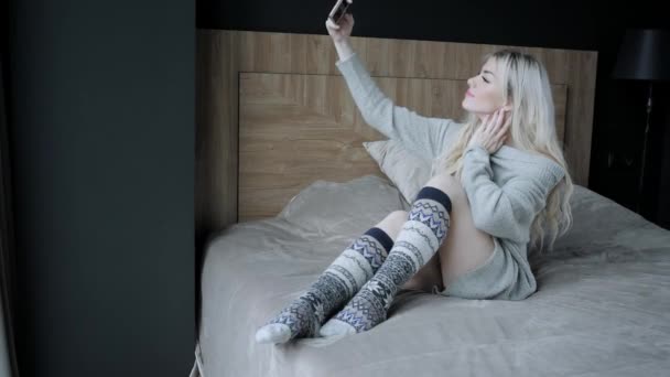 Концепция делает селфи. Мечтательная красивая блондинка сидит на кровати со смартфоном. Девушка улыбается, хорошее настроение. Блоги, интернет, чаты. В теплом уютном свитере и шерстяных носках . — стоковое видео