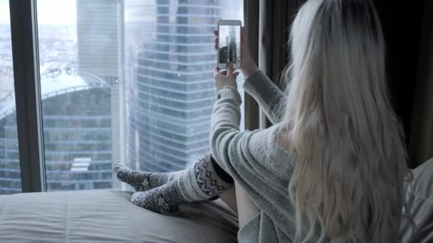夢のような美しい金髪女性がベッドの上に座って窓からスマート フォン表示の画像になりますと ブログ インターネットの閲覧 チャットします 幸せな冬のコンセプト — ストック動画