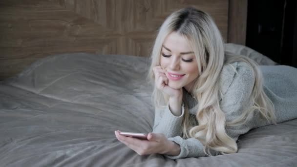 Verträumte schöne blonde Frau auf dem Bett liegend mit Smartphone. Mädchen lächelt, gute Laune. Bloggen, surfen im Internet, chatten. in einem warmen kuscheligen Pullover und Wollsocken. Frohes Winterkonzept. — Stockvideo