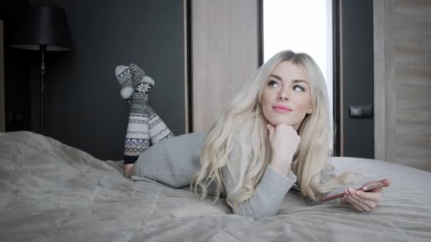 Dreamy Beautiful blonde woman lying on the bed with smart phone. Girl smiles, good mood. Blogging, browsing internet, chatting. In a warm cozy sweater and wool socks. Happy winter concept. — Stock Video