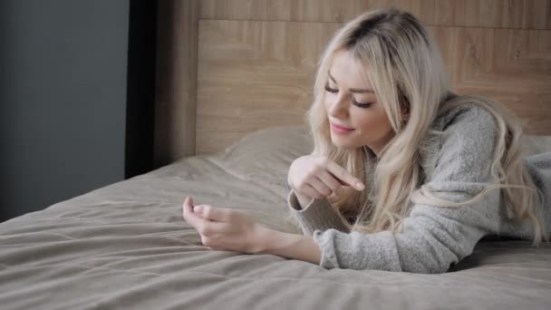 Мечтательная красивая блондинка лежит на кровати со смартфоном. Девушка улыбается, хорошее настроение. Блоги, интернет, чаты. В теплом уютном свитере и шерстяных носках. Счастливая зима . — стоковое видео