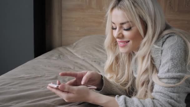 Mooie blonde vrouw liggend op het bed met slimme telefoon. Meisje glimlacht, goed humeur. Bloggen, surfen op internet, chatten. In een warme, knusse trui en wollen sokken. Happy winter concept. — Stockvideo