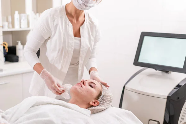 O médico remove o gel do rosto dos pacientes e aplica um creme terapêutico. Fototerapia anti acne. Mulher bonita durante o procedimento de rejuvenescimento da foto. Tratamento da pele da cara na clínica cosmética . — Fotografia de Stock