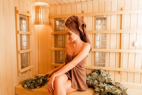 Young woman relaxing in a sauna dressed in a towel. Interior of new Finnish sauna, infrared panels for medical procedures, classic wooden sauna. oak broom
