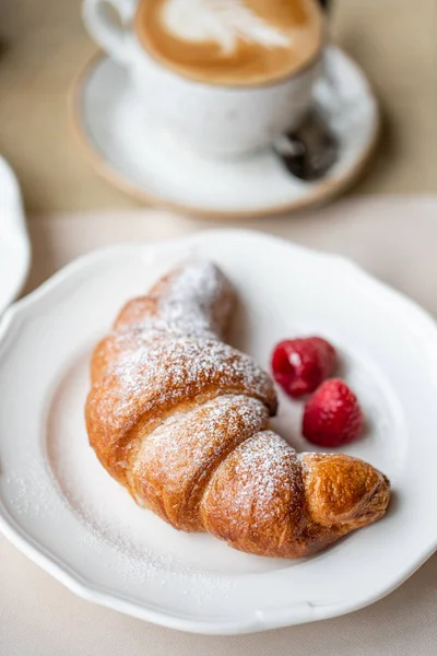 Kaffe cappuccino med croissant på vit platta i restaurangen. Ljus morgon frukost, färska varma bakverk och hallon — Stockfoto