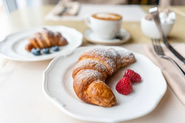 Café capuchino con dos croissant en plato blanco en restaurante. Desayuno ligero por la mañana, pasteles calientes frescos y frambuesas — Foto de Stock