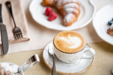 Restoranda beyaz tabakta iki kruvasan ile kahve kapuçino. Işık sabah kahvaltı, taze sıcak hamur işleri ve ahududu