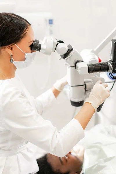 Mujer joven dentista que trata los conductos radiculares con microscopio en la clínica dental. Paciente hombre acostado en silla de dentista con la boca abierta. Concepto de medicina, odontología y salud. Equipos dentales — Foto de Stock