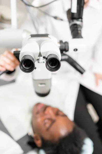 Ocular del microscopio en primer plano. Joven paciente afroamericano en silla en la clínica dental. Medicina, salud, concepto estomatológico. dentista realiza inspección y concluye — Foto de Stock