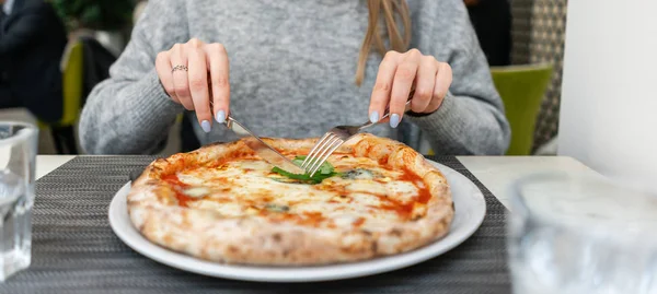 Frau isst mit Messer und Gabel eine Pizza Margherita mit Mozzarella-Tomaten und Basilikum. neapolitanische Pizza aus dem Holzofen. Mittagessen in einem italienischen Restaurant. Tisch neben einem großen Fenster. — Stockfoto