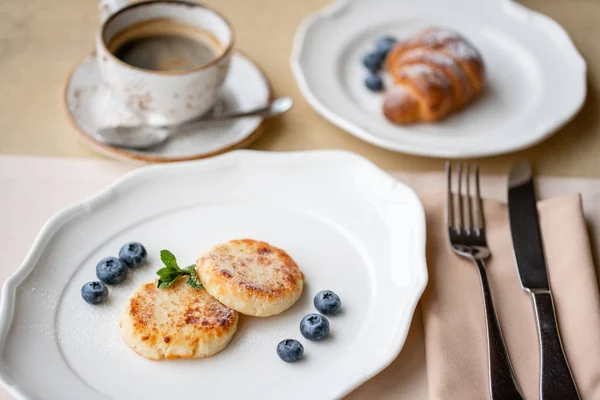 Cheesecake, mirtillo fresco e cappuccino. Frittelle di fiocchi di latte o frittelle di cagliata su piatto bianco. Colazione sana e dietetica all'alba — Foto Stock