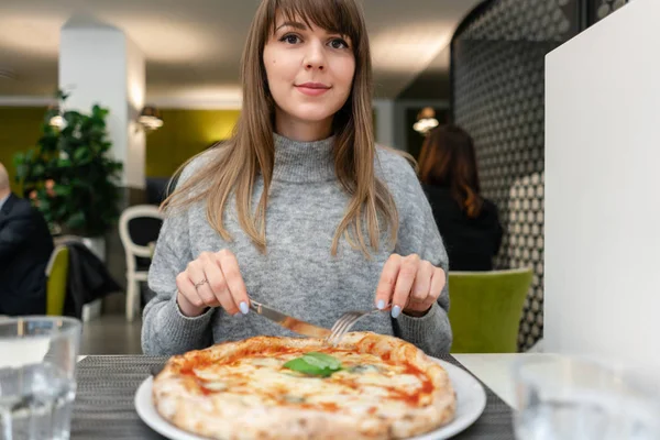 Woman eats with knife and fork a pizza Margherita with mozzarella tomatoes and basil. Neapolitan pizza from wood-burning stove. lunch in an Italian restaurant. Table near to a large window. — Stock Photo, Image