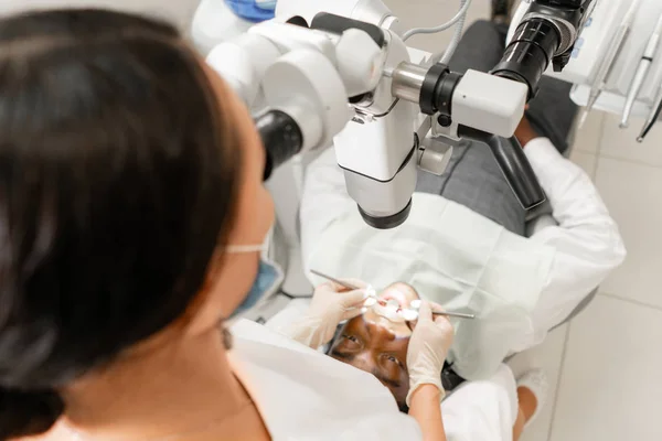 Concéntrate en el microscopio. Mujer joven dentista que trata los conductos radiculares con microscopio en la clínica dental. Hombre paciente acostado en silla de dentista con la boca abierta . —  Fotos de Stock