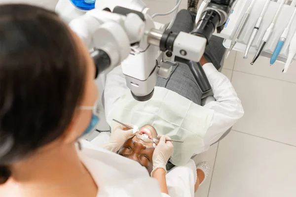 Concéntrate en paciente. Mujer joven dentista que trata los conductos radiculares con microscopio en la clínica dental. Hombre paciente acostado en silla de dentista con la boca abierta . — Foto de Stock