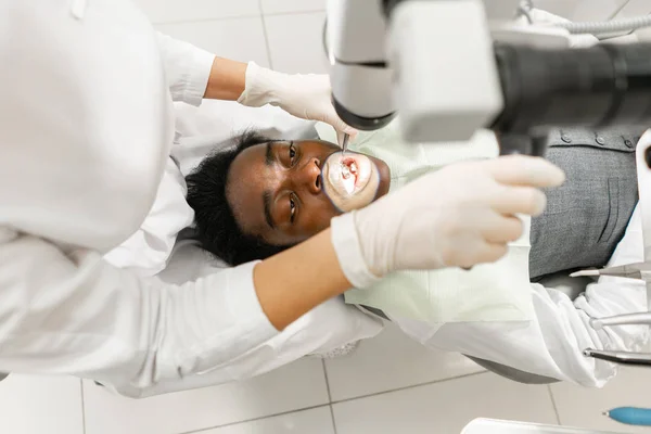 Mujer joven dentista que trata los conductos radiculares con microscopio en la clínica dental. Paciente hombre acostado en silla de dentista con la boca abierta. Concepto de medicina, odontología y salud. Equipos dentales — Foto de Stock