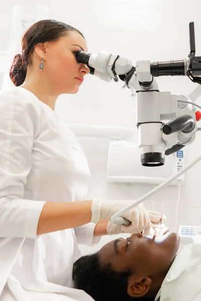 Fiatal nő fogorvos kezelésére gyökércsatornákat használ Mikroszkóp a fogászati klinikán. Férfi beteg feküdt a fogorvos szék, nyitott szájjal. Orvosi, fogorvosi és egészségügyi koncepció. Fogászati berendezés — Stock Fotó