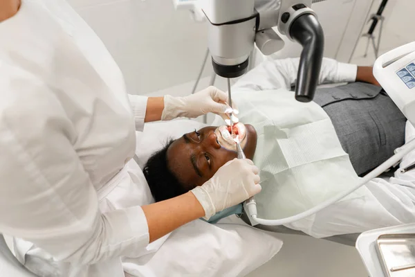 Mujer joven dentista que trata los conductos radiculares con microscopio en la clínica dental. Paciente hombre acostado en silla de dentista con la boca abierta. Concepto de medicina, odontología y salud. Equipos dentales — Foto de Stock