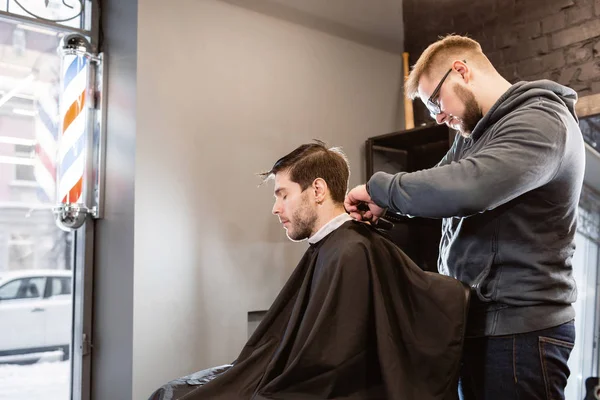Master cuts hair and beard of men in the barbershop, hairdresser makes hairstyle for a young man. Barber work with clipper machine in barbershop.
