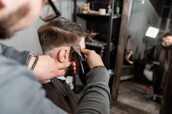 Mester csökkenti a haját és szakállát a férfiak a fodrászatot, fodrász teszi a frizura egy fiatal férfi. Fodrász munka Clipper gép Barbershop. — Stock Fotó