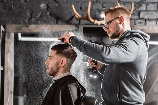 Barber sprays clean water on head in barbershop. Professional trimmer tool cuts beard and hair on young guy in barber shop salon.