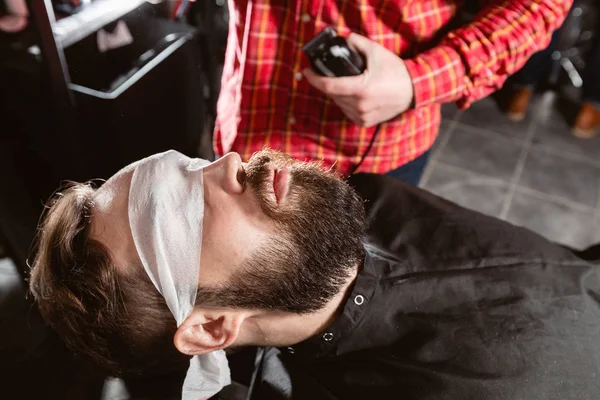 Парикмахер работает с клиппером в парикмахерской. Профессиональный триммер стрижет бороду и волосы молодого парня в парикмахерской . — стоковое фото