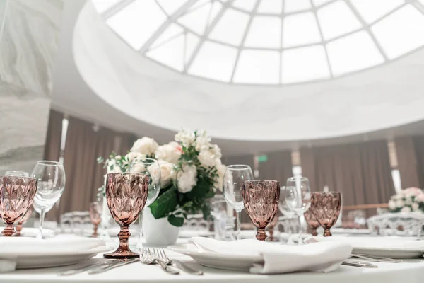 Wine glasses on round Banquet table served. Interior of restaurant for wedding dinner, ready for guests. Decorated with floral arrangement. Dishes, wine glasses and napkins. Catering concept.