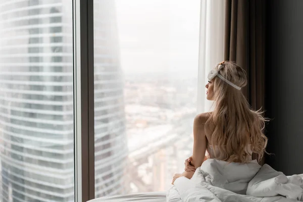 Morning in hotel room. Young woman sits on comfortable bed in mask for sleeping on head. Window of a skyscraper on background.