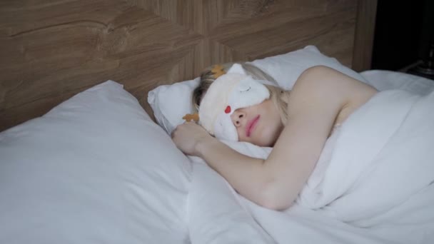 Young woman sleep on comfortable bed in a mask for sleeping. Blindfold on eye. Morning in hotel room. White pillow and blanket — 비디오