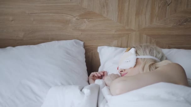 Young woman sleep on comfortable bed in a mask for sleeping. Blindfold on eye. Morning in hotel room. White pillow and blanket — Stock Video