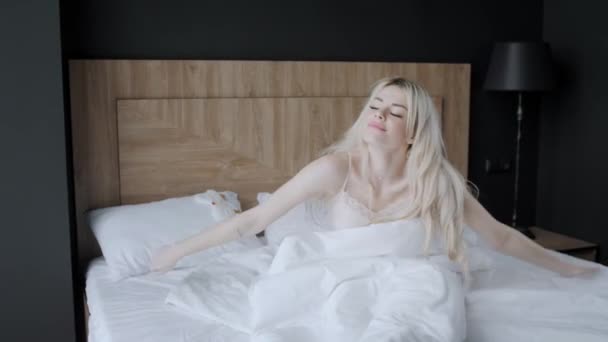 Morning in hotel room. Young woman sits on comfortable bed in mask for sleeping on head. Window of a skyscraper on background. — Stock Video